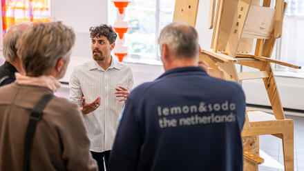 Een jonge kunstenaar staat bij een kunstwerk van op elkaar gestapelde houten stoelen. Hij spreekt een groep bezoekers toe.