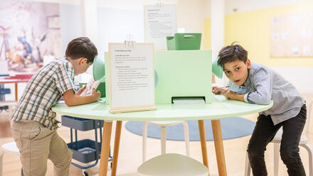 Zwei Jungen stehen an einem runden Tisch. Sie halten ihr Ohr an einen Trichter und lauschen auf etwas. Der Raum ist bunt und sie schauen neugierig.