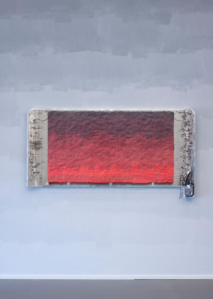 Een kunstwerk hangt aan de muur. De zijkanten zijn grijs, met allerlei elektrische draden die eruit steken. Het middenvlak bestaat uit verschillende tinten rood.
