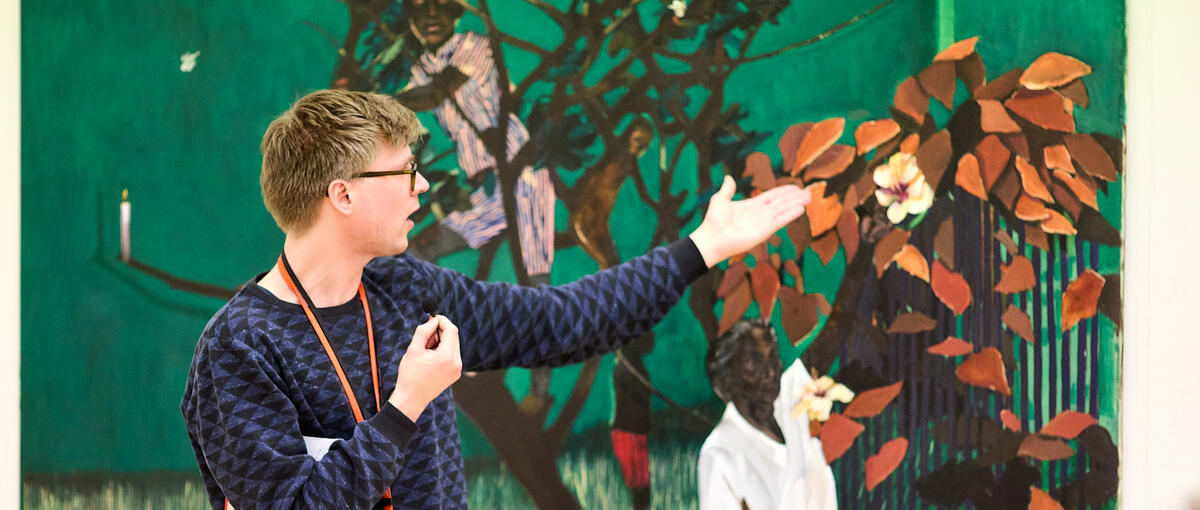A male museum docent stands in front of a green painting. He is pointing to the right and talking. In the foreground, you can see the backs of listening students.