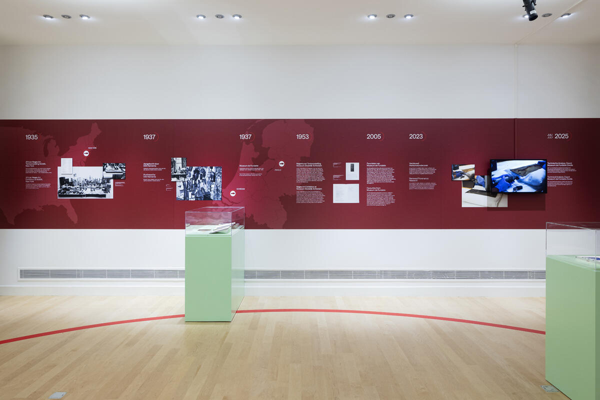 Een muur van het museum met daarop een rode strook op de muur. Op deze strook staat allemaal informatie en foto's van het herkomstonderzoek van Back to Benin.