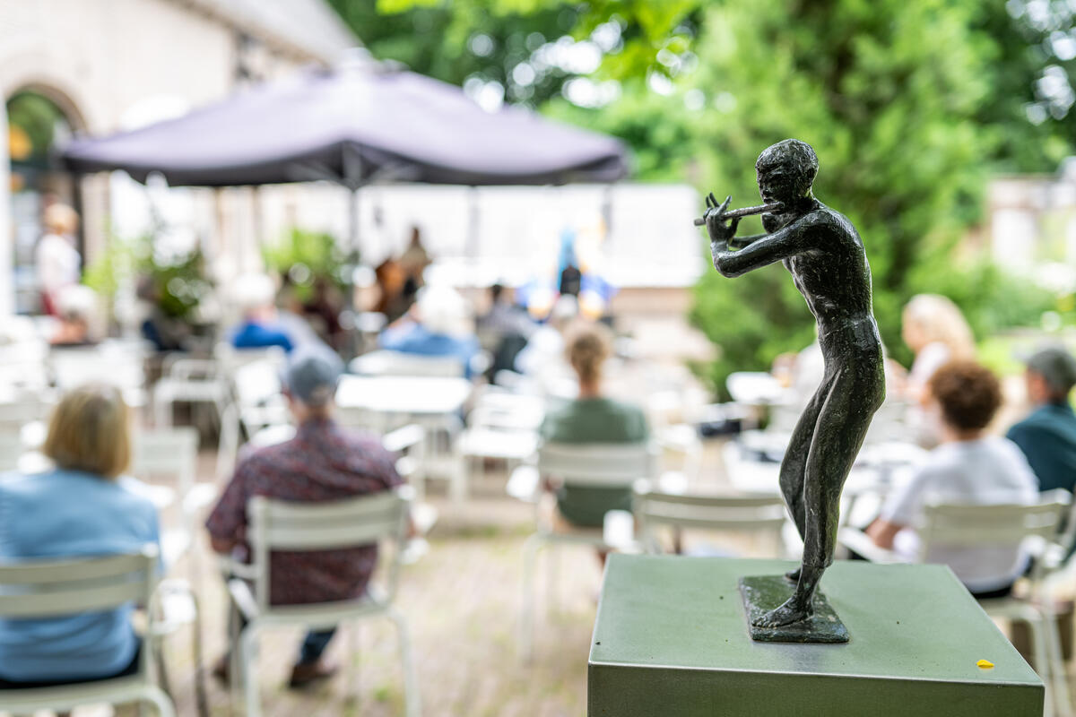 Een klein sculptuur van een man die een dwarsfluit speelt staat op de voorgrond. Op de achtergrond is het terras van Kasteel het Nijenhuis zichtbaar.