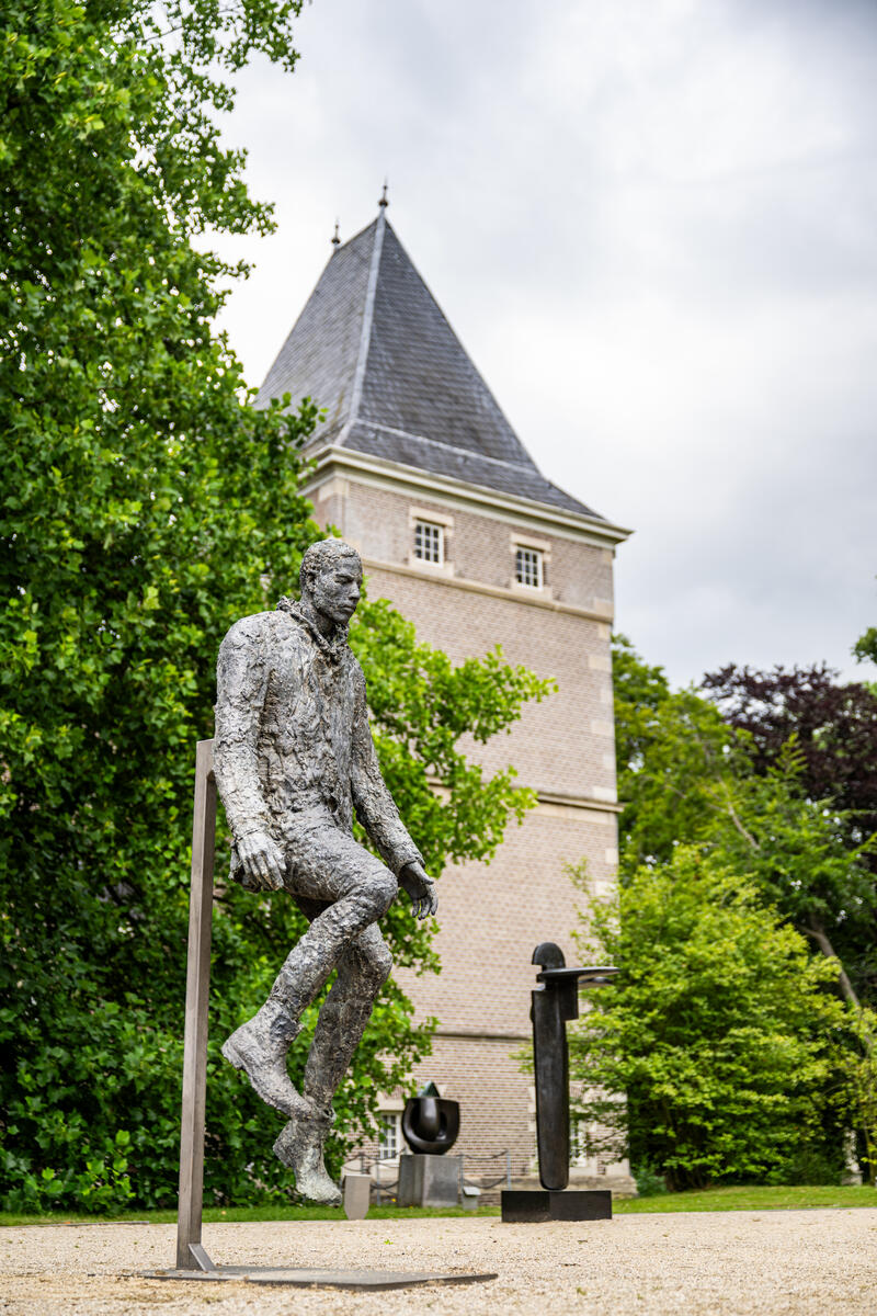 Een standbeeld van een man staat op de voorgront. Achter het standbeeld staat een grote boom en is een toren van Kasteel het Nijenhuis zichtbaar.