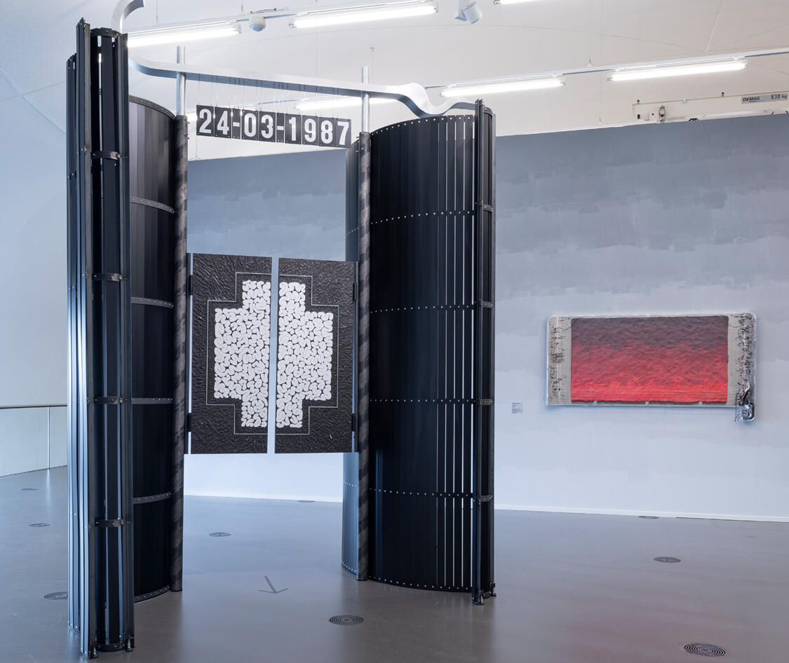 Een ruime museumzaal met links een grote zwarte installatie van gebogen panelen. In het midden hangt een zwart-wit kruisvormig kunstwerk met daarboven de datum 24-03-1987. Rechts aan de muur hangt een langwerpig kunstwerk met een rode gloed.