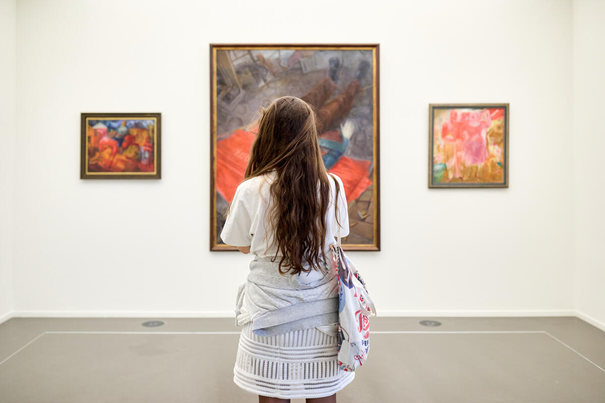 Een jonge vrouw staat voor een groot schilderij. Ze heeft een trui om haar middel geslagen. Verder in de ruimte leeg.