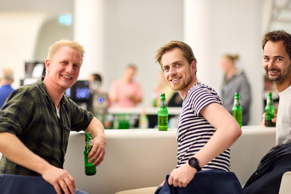 Drie jonge bezoekers zitten bij de bar van het museum met een biertje. Ze draaien zich om naar de camera en lachen.