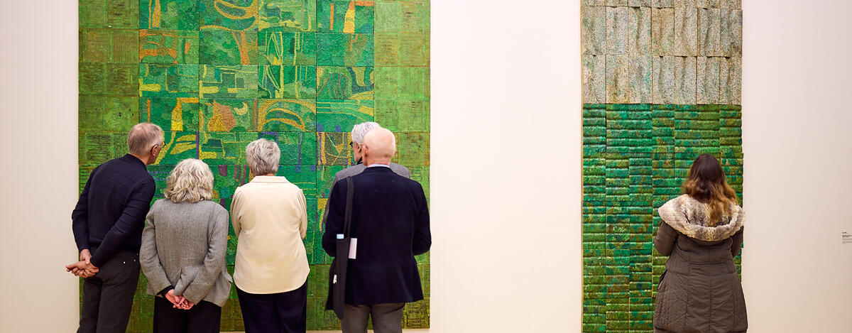 Een groep bezoekers staat dicht bij groene kunstwerken die aan de muur van het museum hangen. 