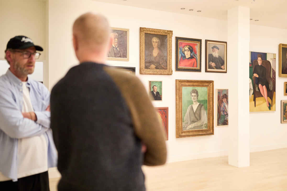 Twee mannelijke bezoekers praten met elkaar in een zaal waar allemaal portretten van vrouwen aan de muur hangen. De mannen zijn onscherp.