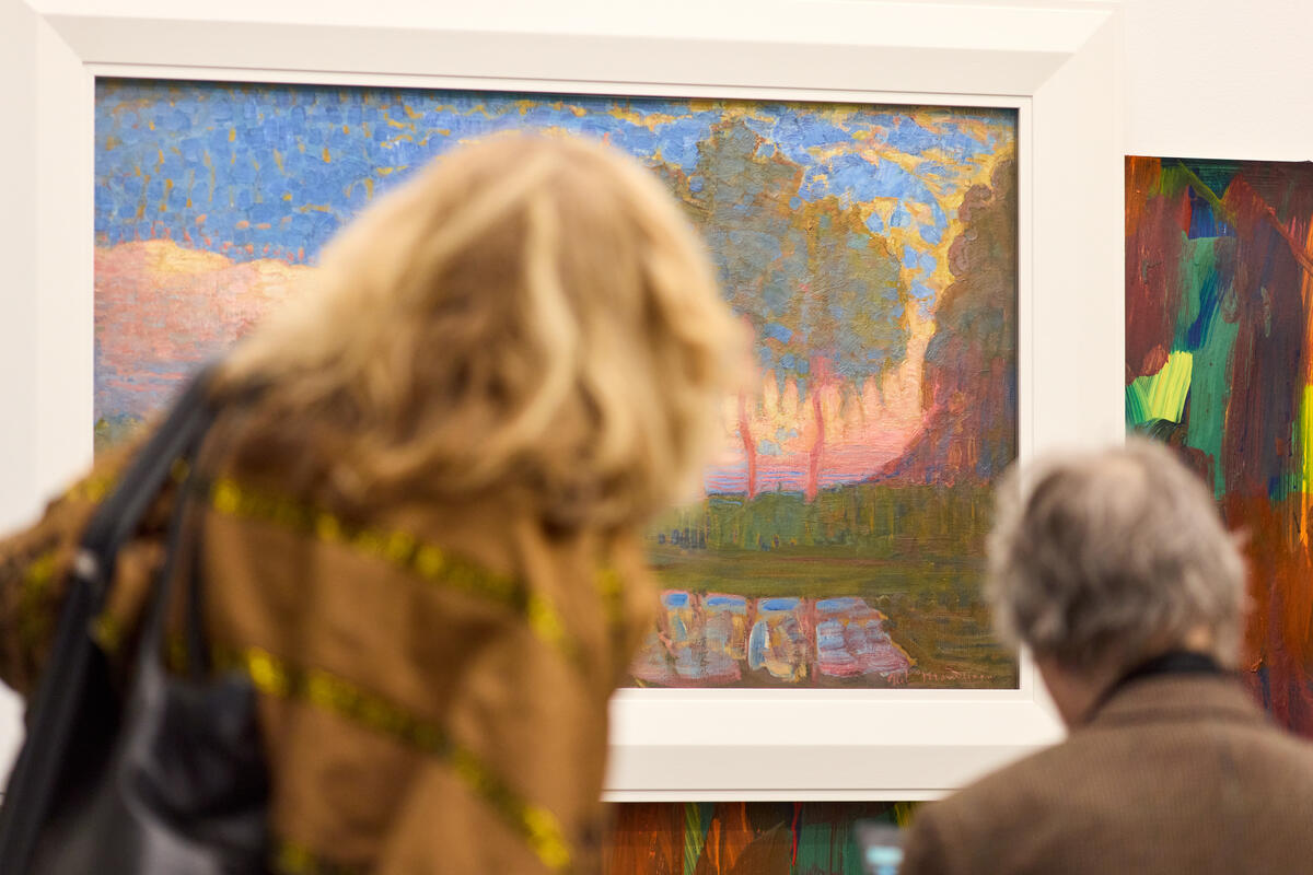 Een man en een vrouw kijken naar een impressionalistisch schilderij met warme kleuren van een landschap.
