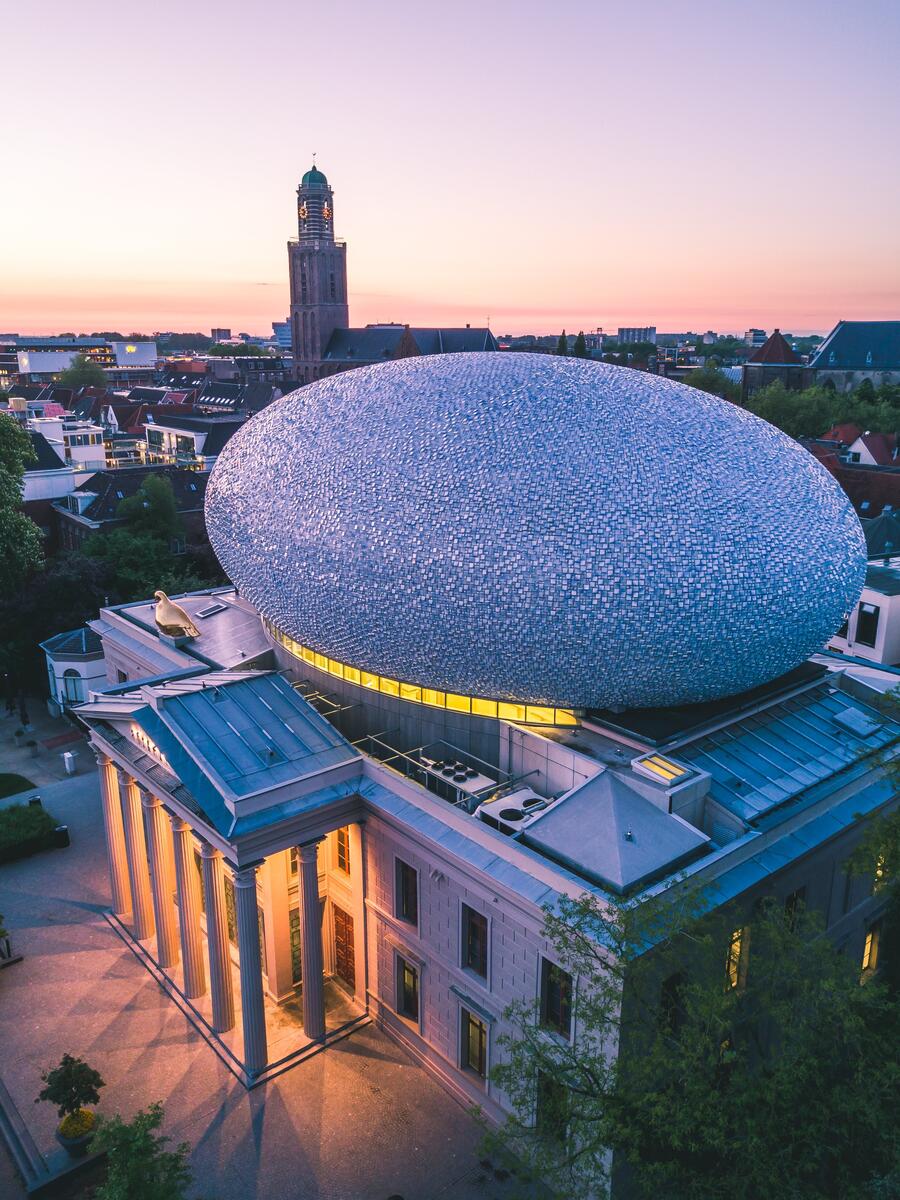 An aerial view of Museum De Fundatie in Zwolle. It is dusk and the lights beneath the columns cast a warm glow. The sky is pale pink.