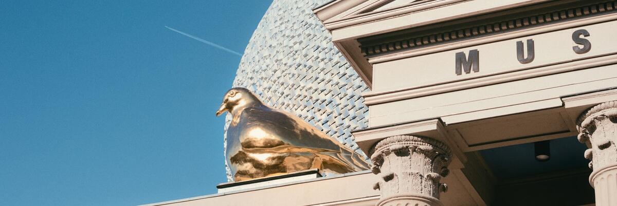 Een detail van het beeld de gouden vredesduif die op het dak van museum de FUndatie zit. De lichtblauwe opbouw en pilaren zijn zichtbaar aan rechterkant van de foto.