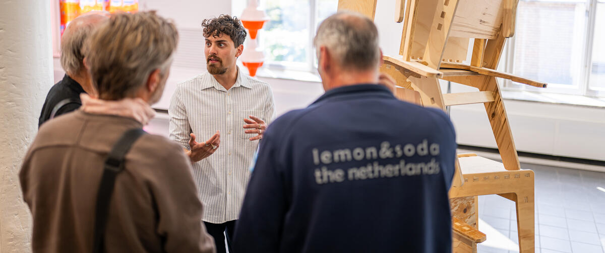 A young artist stands by an artwork of stacked wooden chairs. He addresses a group of visitors.
