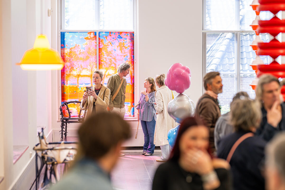 Bezoekers staan in een witte ruimte met kleurrijke kunstvoorwerpen. Een vrouw maakt een foto van een kunstwerk.