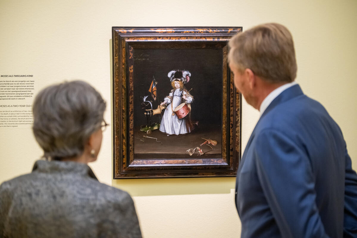 Koning Willem-Alexander staat met een vrouw voor een van de bekende werken van Gesina ter Borch, Portret van Moses ter Borch als tweejarig kind. Op het schilderij is broertje Moses te zien in een jurk met wit schort en eenhoed met veren die scherp afsteken tegen de donkere achtergrond. De jongen draagt een trom en trommelstokken.