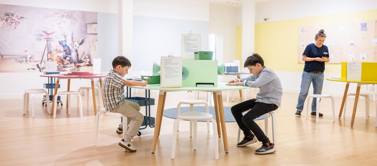 Twee jongens zitten in De Studio aan een ronde tafel te tekenen. Een vrouw staat bij een andere tafel.