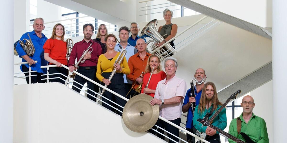 Muzikanten staan in een wit trappenhuis. Ze dragen allemaal verschillende kleuren shirts en houden hun instrumenten vast voor de foto. 