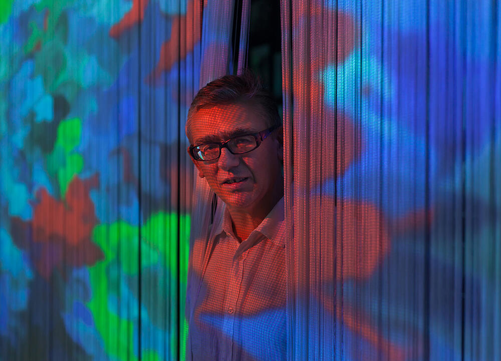 Een portret van Miguel Chevalier. Hij steekt zijn hoofd door een gordijn van dunne touwtjes waar een kleurrijk digitaal kunstwerk op geprojecteerd is. 