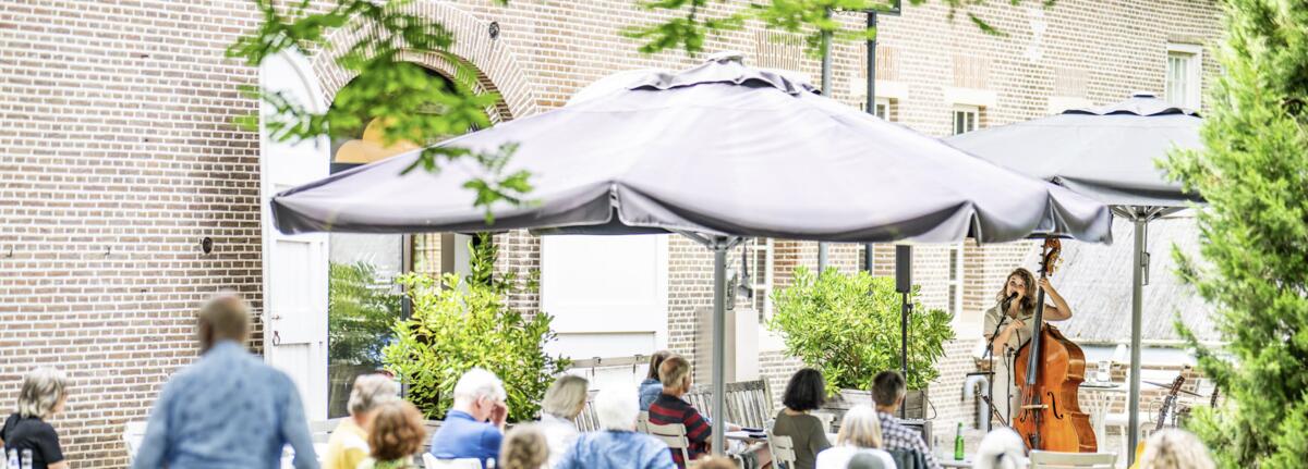 Mensen zitten op een terras te luisteren naar een vrouw die contrabas speelt en zingt. 