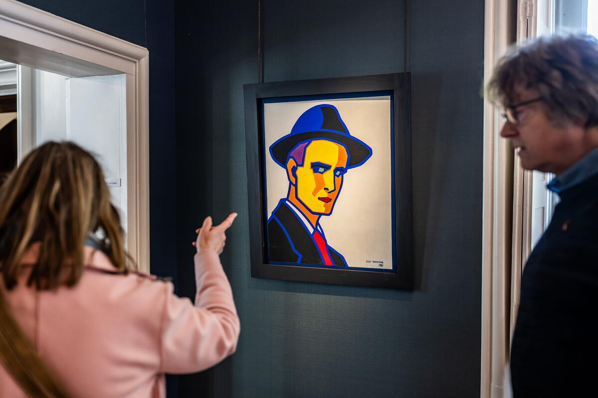 Een bezoeker in een roze jas wijst naar een schilderij van een man met een hoed op die schuin naar haar lijkt terug te staren.