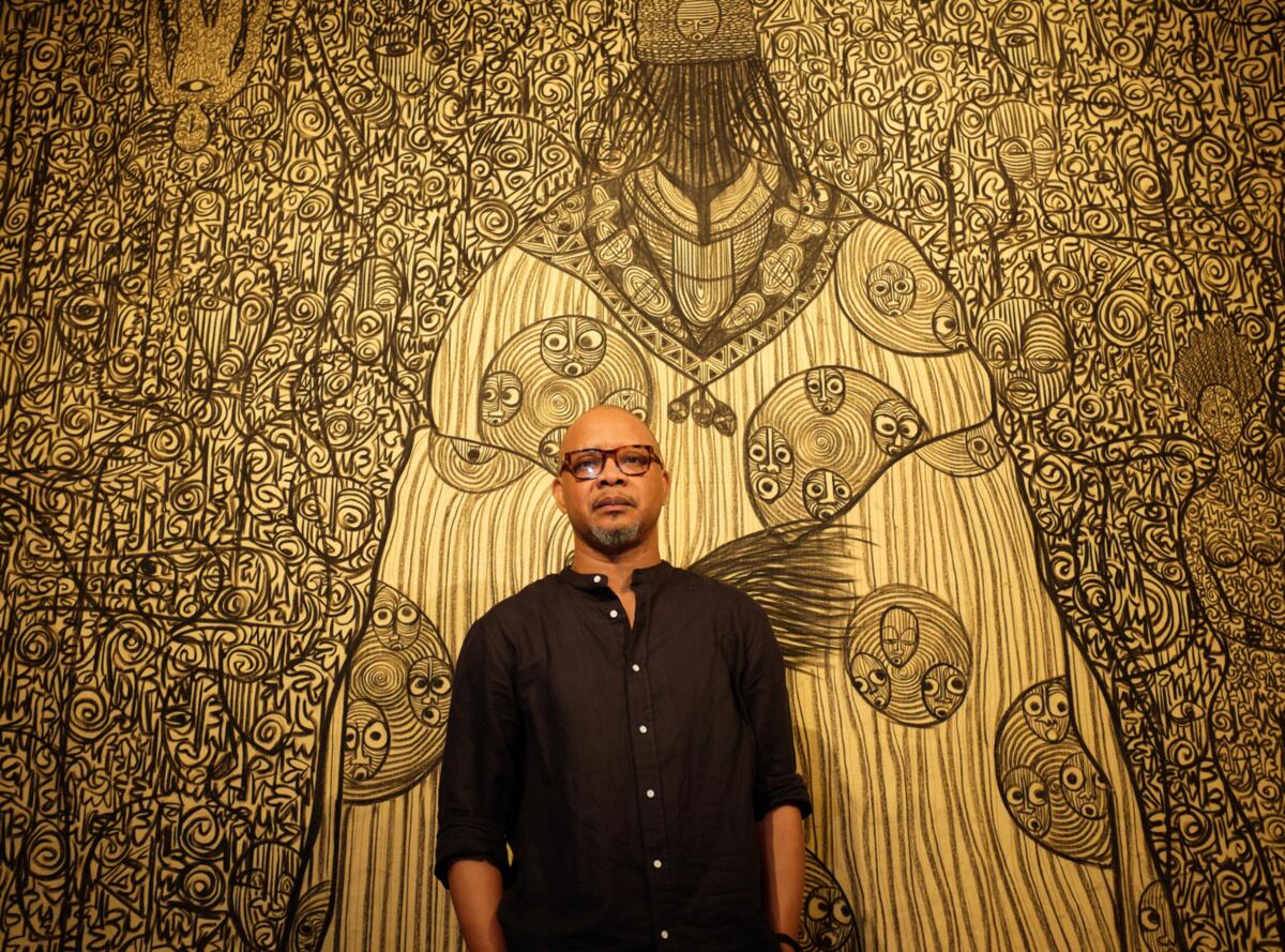 Portret van kunstenaar Victor Ehikhamenor. Achter hem hangt muurvullende tekening in geel- en bruintinten.