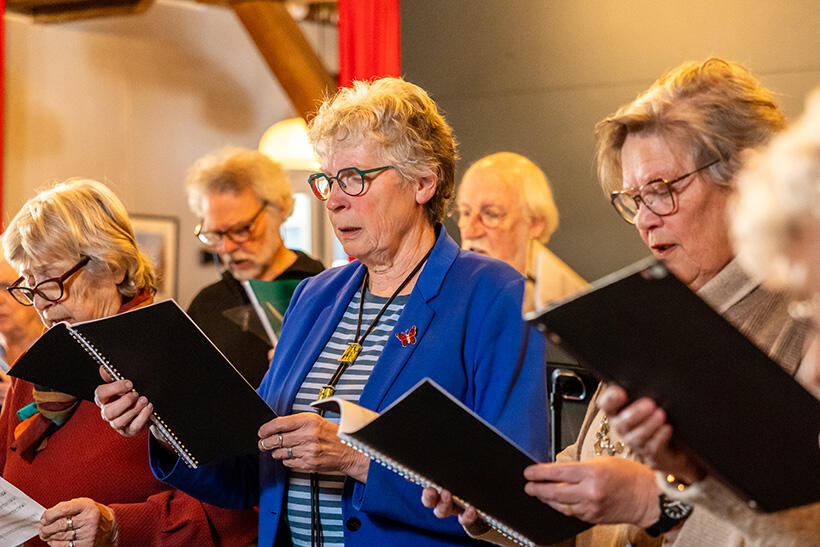 Deelnemers van Heino Vocaal staan te zingen. Ze lezen bladmuziek.