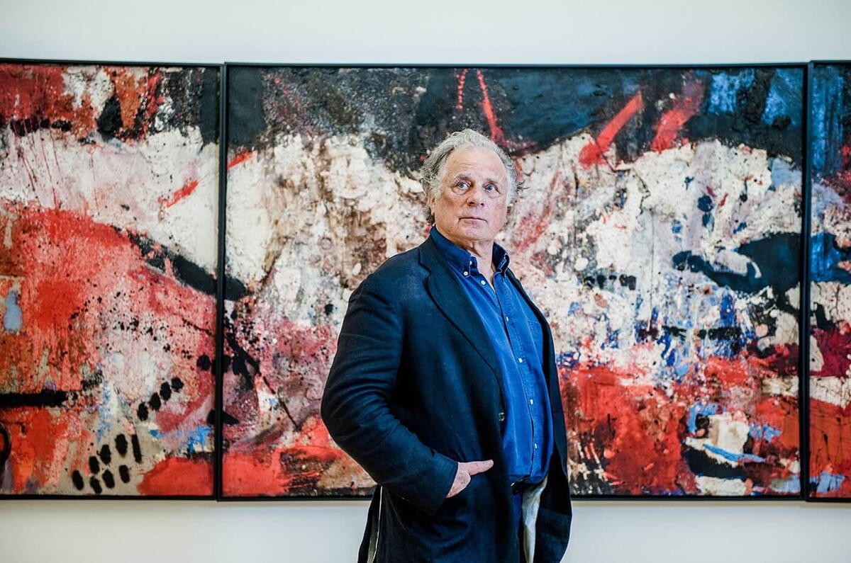 Een oudere Jan Cremer staat voor een groot schilderij in rood en donkerblauw. Cremer draagt zelf een donkerblauw pak en kijkt naar de verte.