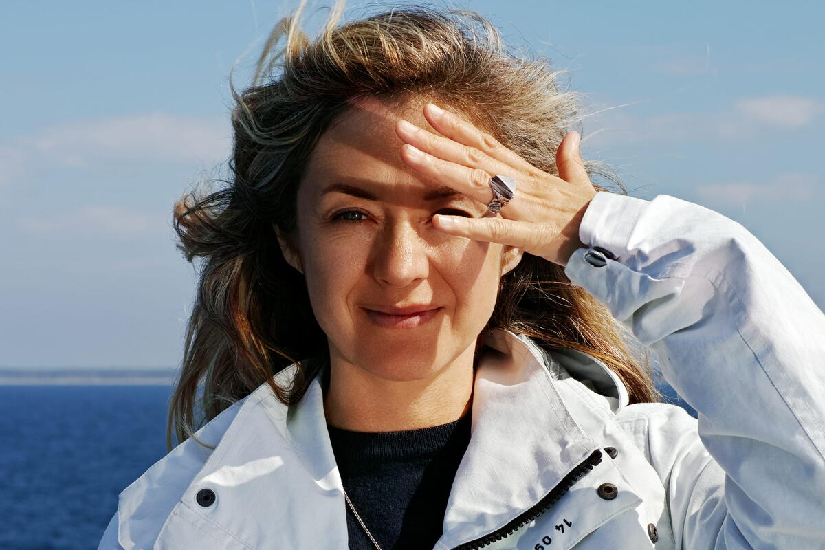 De kunstenares staat voor de zee. Ze heeft een hand voor haar gezicht en glimlacht. Haar haren zijn naar achter geblazen en je ziet het blauw van de zee.