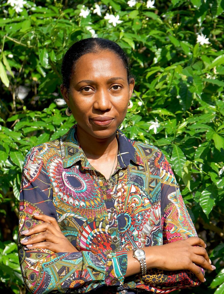 Portretfoto van Taiye Idahor. Ze draagt een gekleurde blouse en staat buiten voor een groene heg.