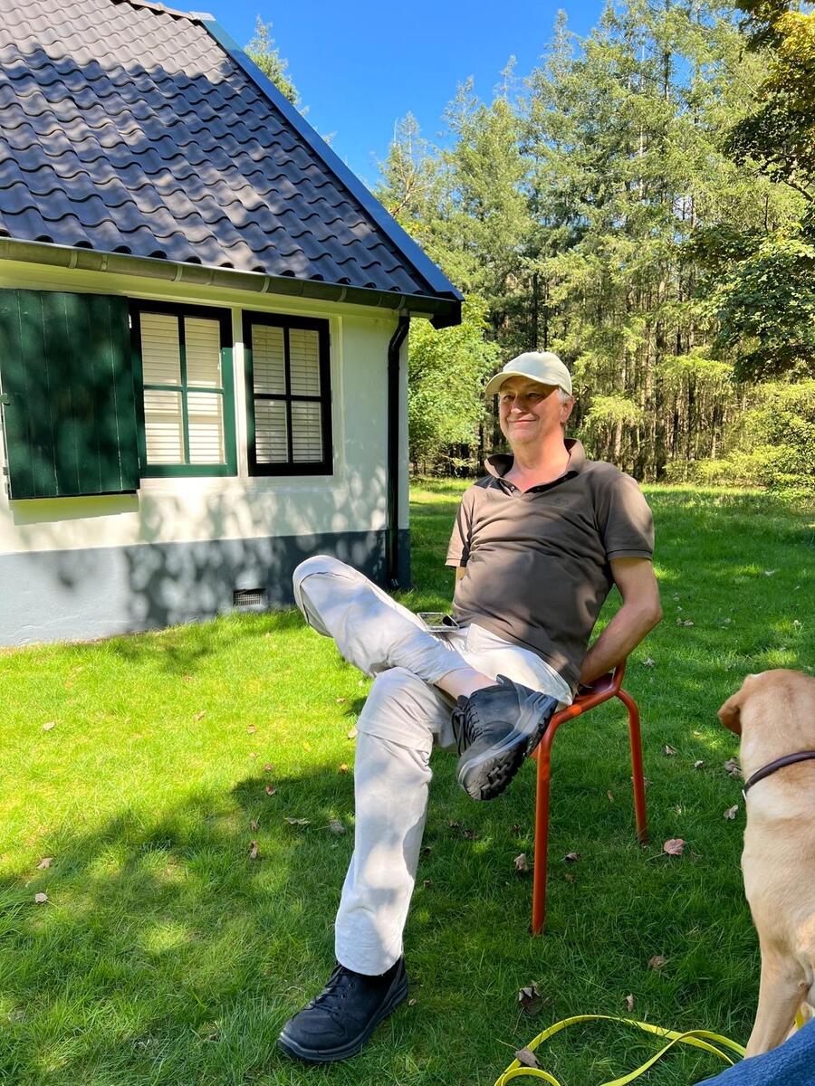 Foto van kunstenaar Q.S. Serafijn. Hij zit ontspannen op een rode stoel voor een huisje in het bos. 