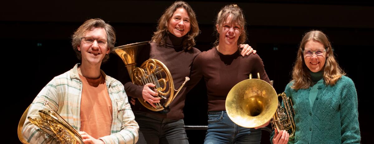 Een groepsfoto van vier muzikanten die lachen naar de camera met hun blaasinstrumenten naast hen.