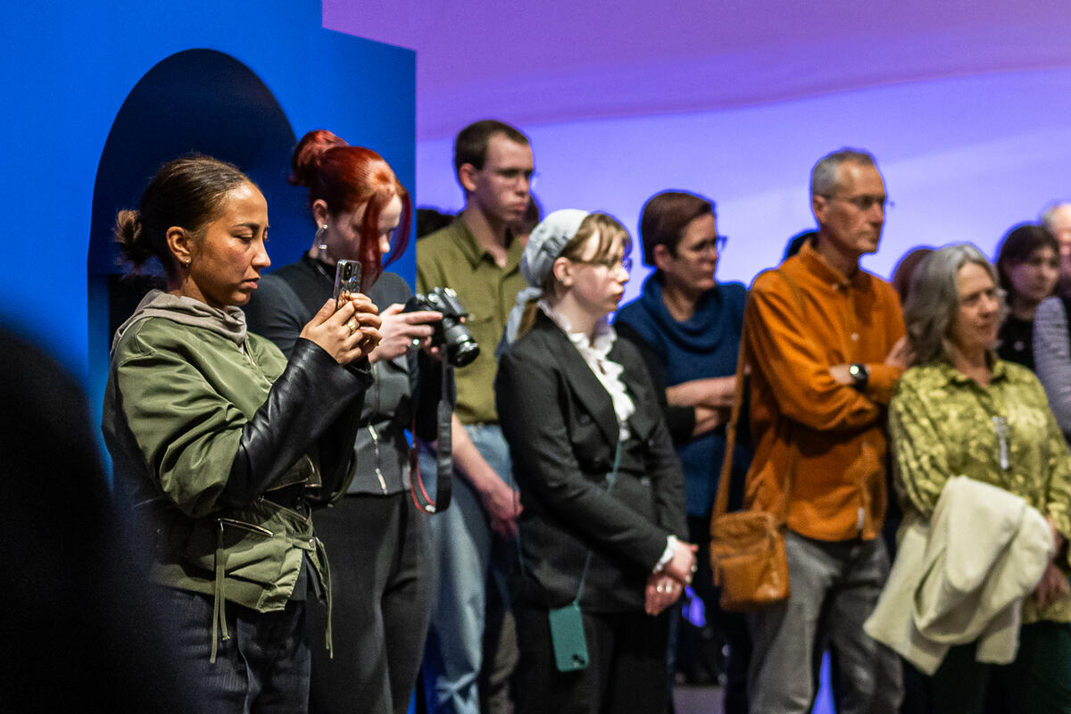Groep bezoekers staat in een museumzaal met blauw-paars licht te luisteren en te kijken. Vooraan maken twee personen foto's met een telefoon en camera.