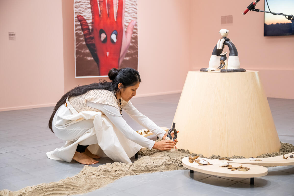 Cristina Flores Pescorán bij haar eigen kunstwerk. Het kunstwerk bestaat uit verschillende materialen zoals zand, hout en beeldhouw kunstwerken. De kleuren zijn licht en naturel.