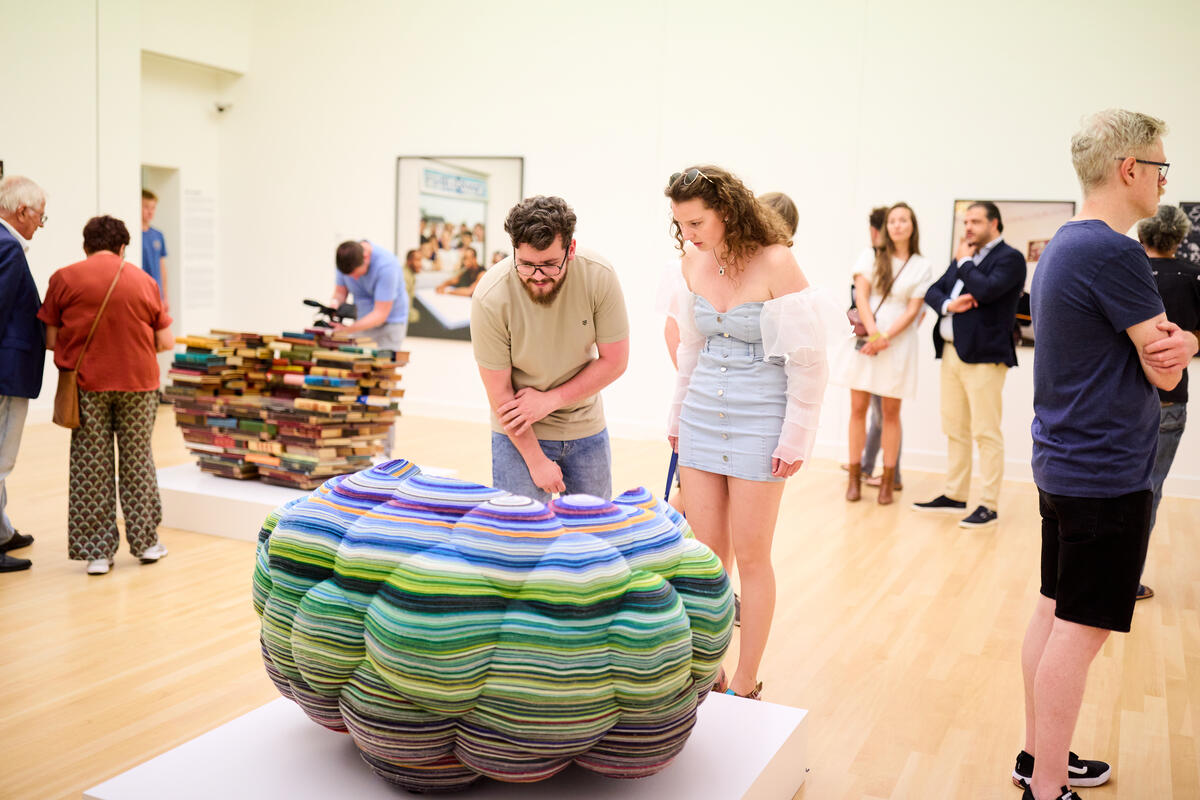 Bezoekers bekijken kunstwerken in een lichte museumzaal, waaronder een grote, kleurrijke, gelaagde sculptuur op een sokkel en een stapel boeken op de achtergrond.