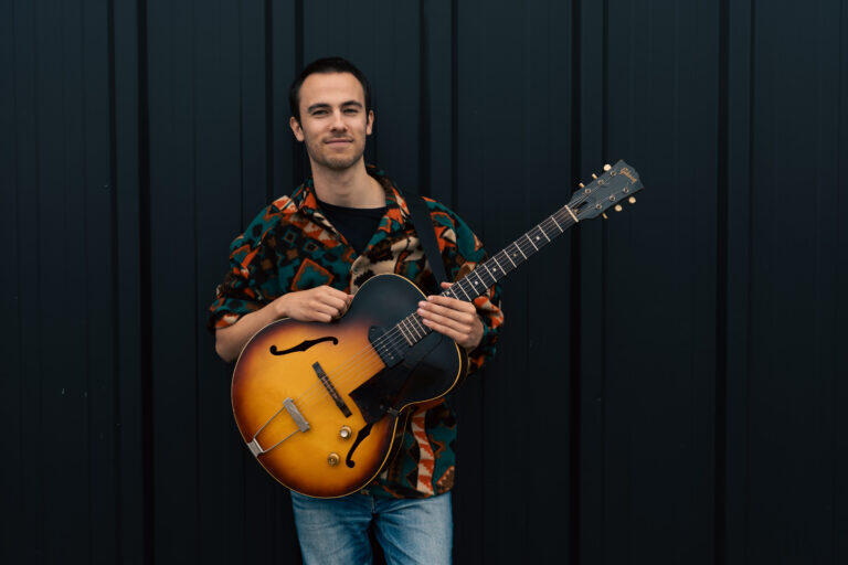Foto van Jim van de Klundert samen met zijn gitaar voor een zwarte achtergrond