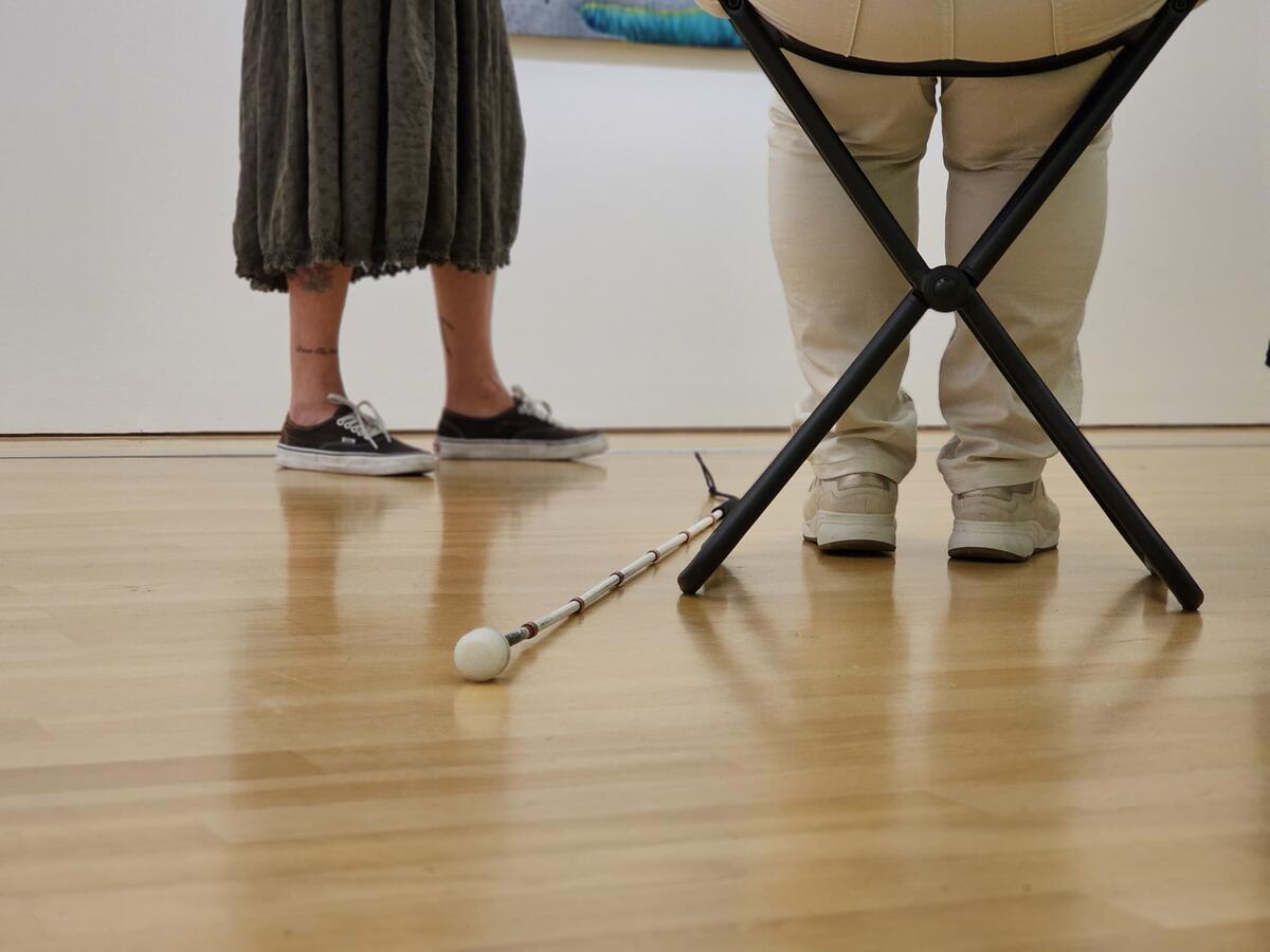 Een blindegeleidestok ligt op de vloer van het museum. De benen van een vrouw zijn zichtbaar. Ze zit op een kruk.