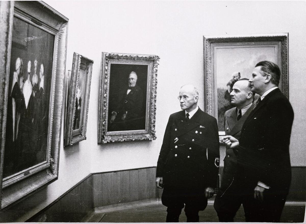 Een jonge Dirk Hannema staat met twee mannen in pak voor een muur met schilderijen. De foto is in zwart-en-wit.