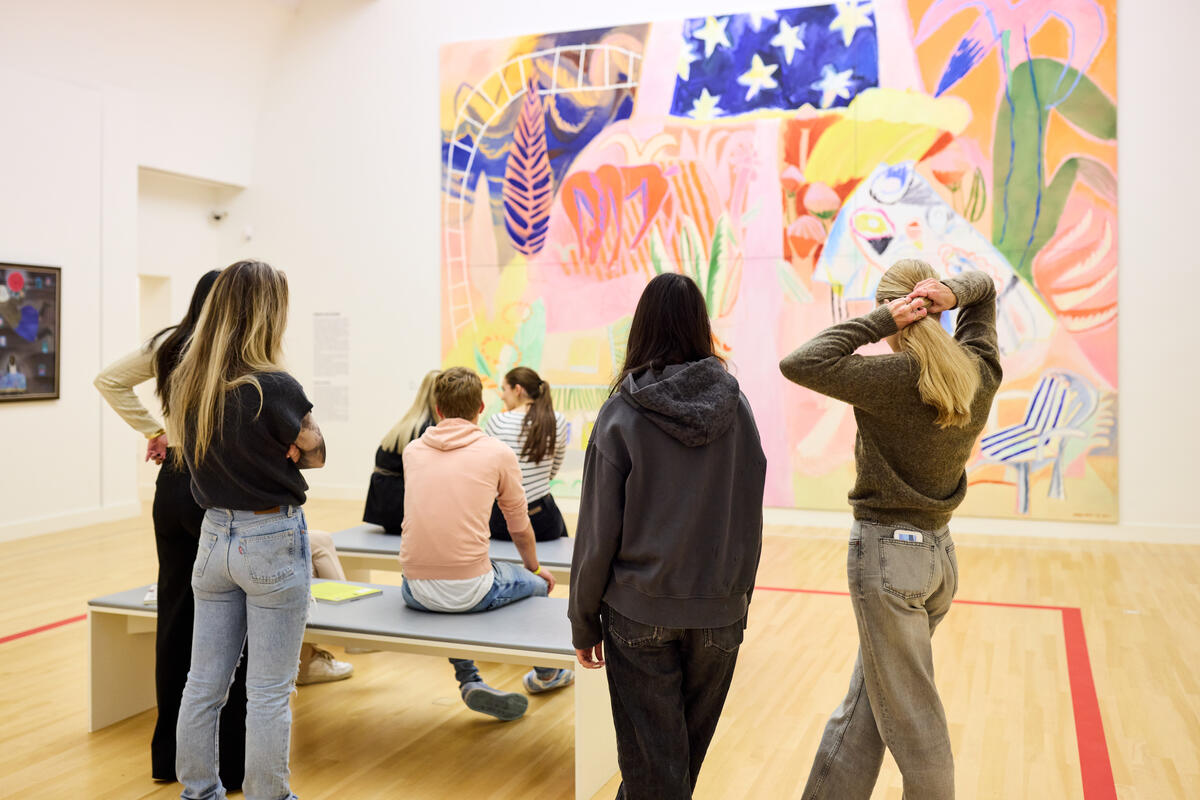 Scholieren staan en zitten in een zaal van het museum. Ze kijken naar een groot kleurrijk schilderij met veel roze vlakken.