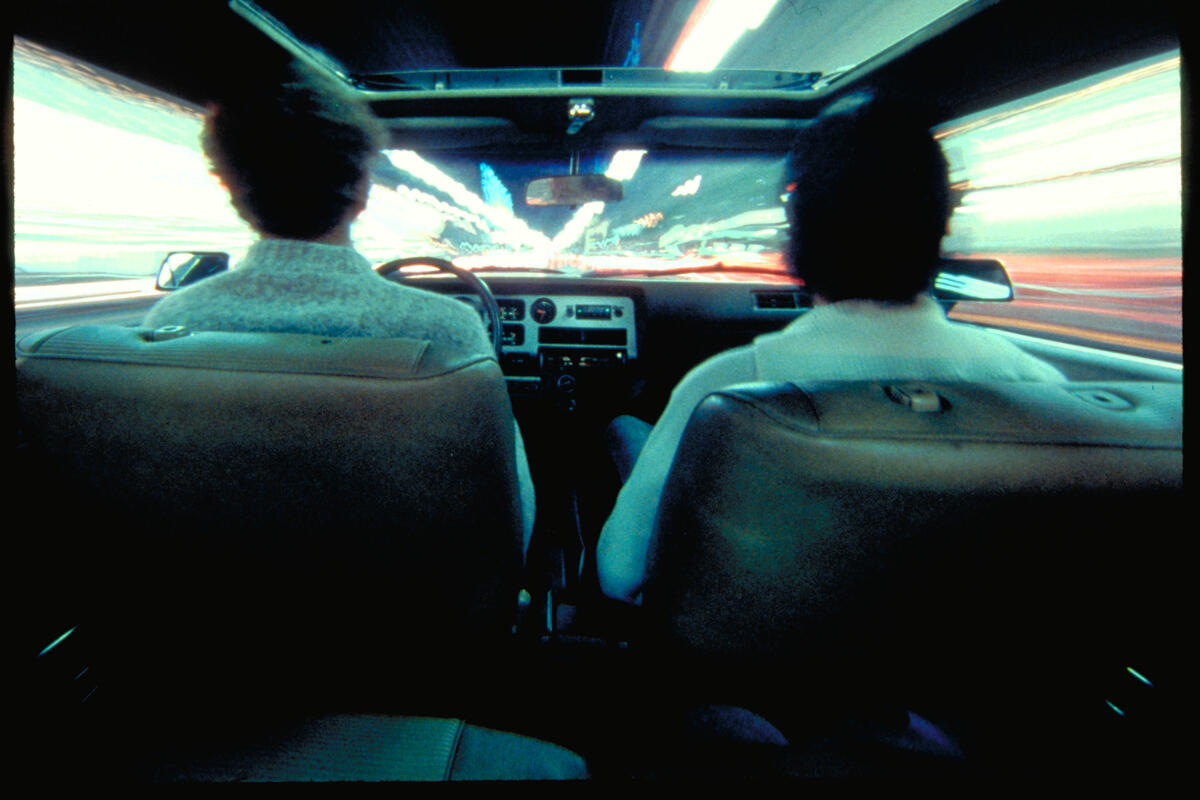 Filmstill aus &bdquo;Koyaanisqatsi&ldquo;, der zwei Personen in einem Auto vom R&uuml;cksitz aus zeigt