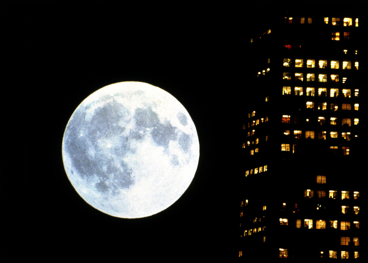 Filmstill aus &bdquo;Koyaanisqatsi&ldquo;, der einen Vollmond neben einem Wohnblock zeigt