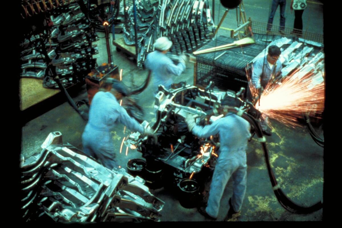Filmstill aus &bdquo;Koyaanisqatsi&ldquo;, der Menschen zeigt, die an einem Auto arbeiten
