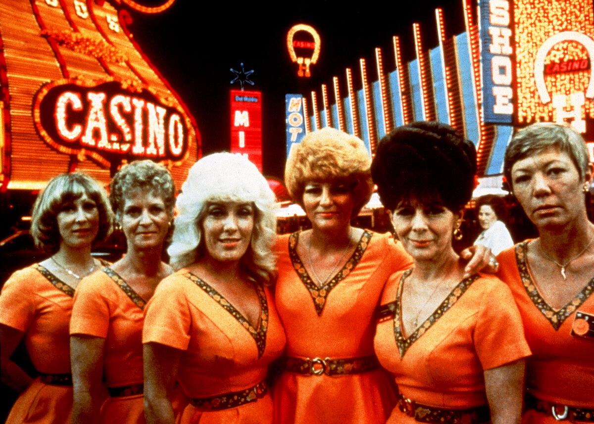 Ein Filmstill aus &bdquo;Koyaanisqatsi&ldquo;, der sechs Frauen zeigt, die in identischen orange-gestreiften Kleidern vor einem orange beleuchteten Casino stehen