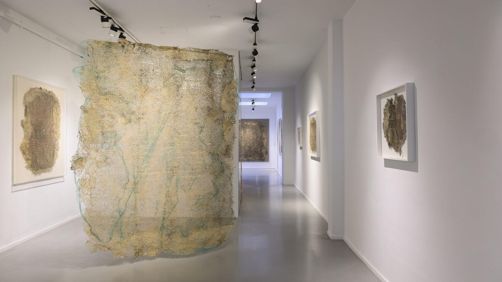 Een atelier waarbij aan de muren lijsten hangen met kunst gemaakt van plantenwortels. In het midden van de ruimte hangt een doek gemaakt van textiel en organisch materiaal met blauwe wol er doorheen verwerkt.