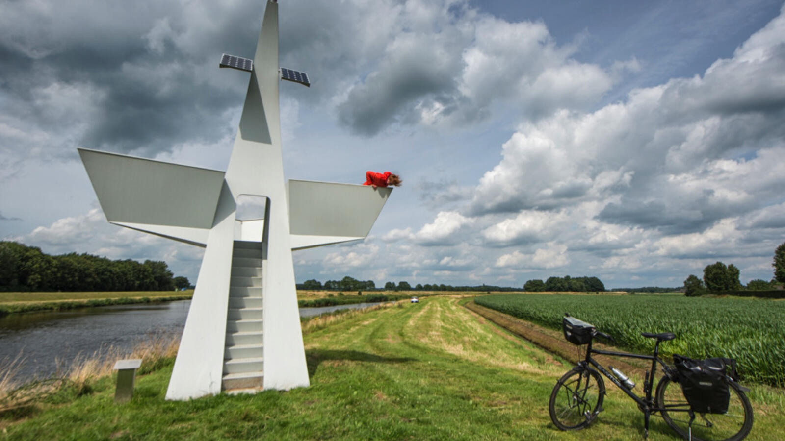 Een metershoog wit kunstwerk staat in een wijd landschap. Het kunstwerk dient ook als uitkijktoren en heeft de vorm van een zeil van een schip. Een vrouw met rode jas hangt over de balustrade. Haar fiets staat in het gras beneden.