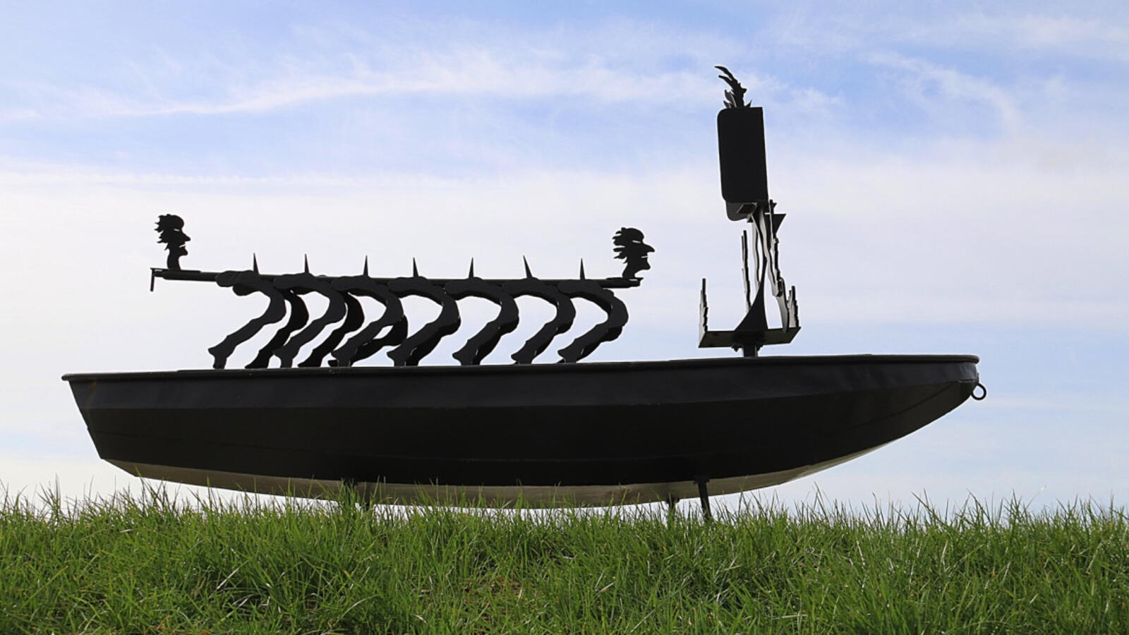 Een kunstwerk in de vorm van een ijzeren boot met daarboven abstracte vormen, waaronder twee silhouetten van gezichten, staat in het gras met een blauwe lucht erachter.