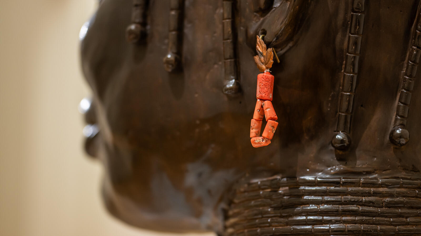 Een detail van een bronzen standbeeld van een Nigeriaanse vrouw met kettingen om haar nek. Ze draagt een oranje oorbel van kralen met een bladmotief aan de bovenkant.