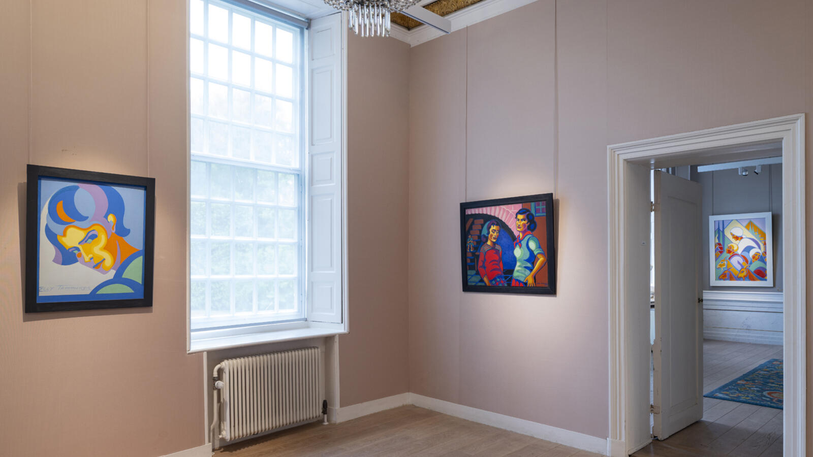 Kleurrijke schilderijen van Tamminga hangen in een lichtroze ruimte. Aan het plafond hangt een kroonluchter.