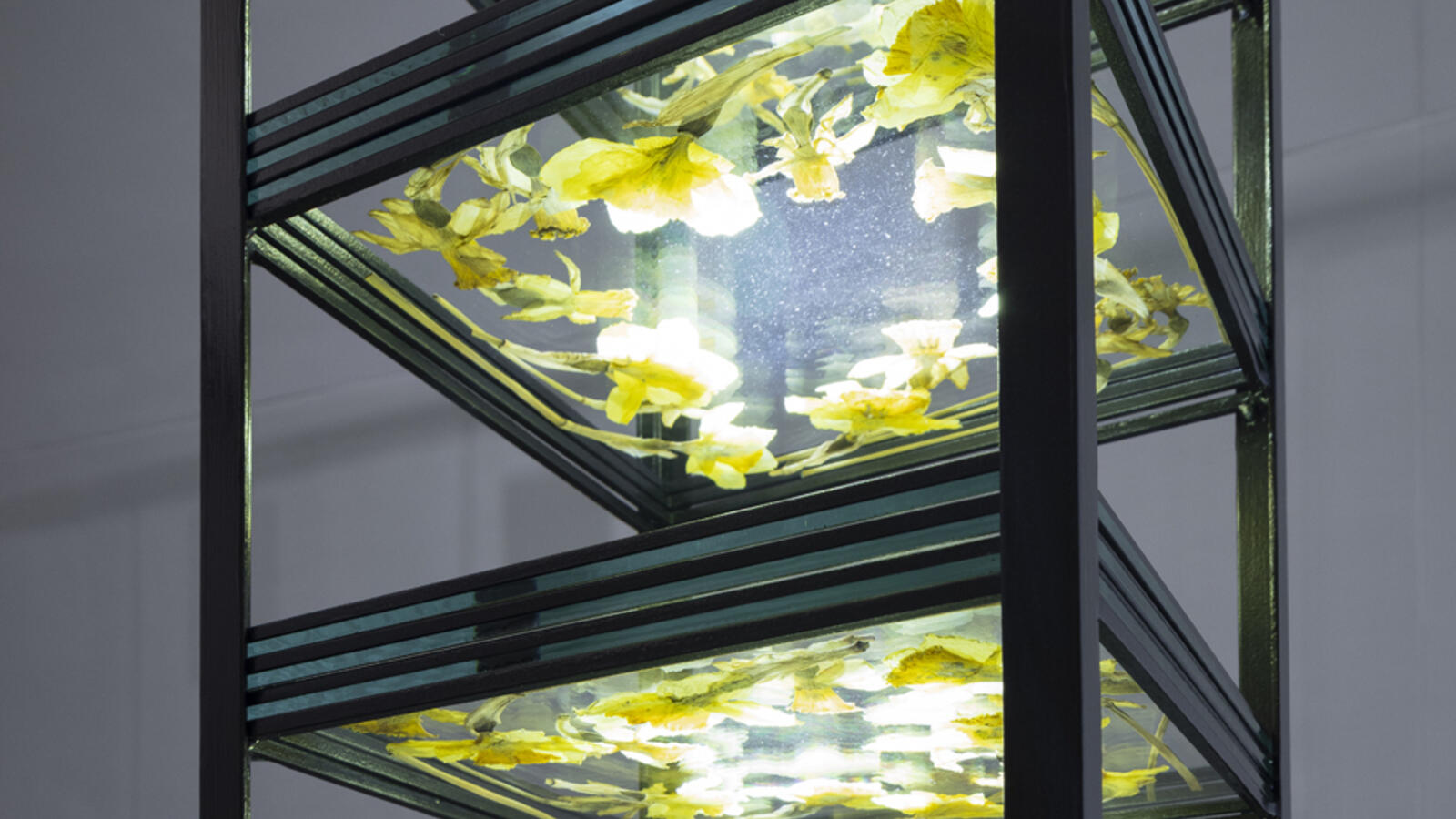 Een metalen toren met afgewisseld glazen laten en lege ruimte. Per laag liggen drie glazen platen op elkaar met gele bloemen ertussen.