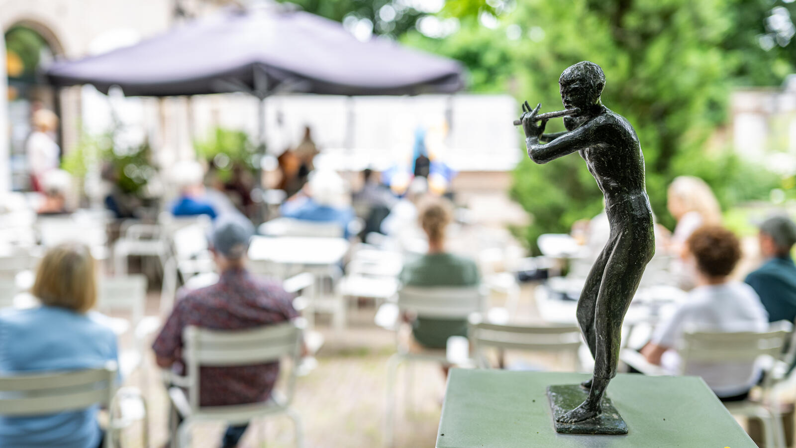 Een klein sculptuur van een man die een dwarsfluit speelt staat op de voorgrond. Op de achtergrond is het terras van Kasteel het Nijenhuis zichtbaar.