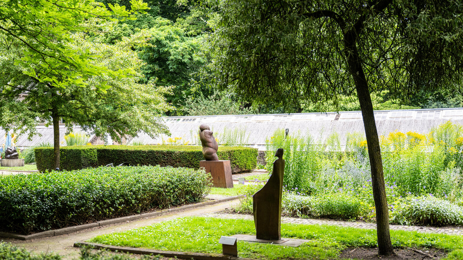 A view of a garden with hedges in the sculpture garden. Two small sculptures stand on the grass, surrounded by trees.