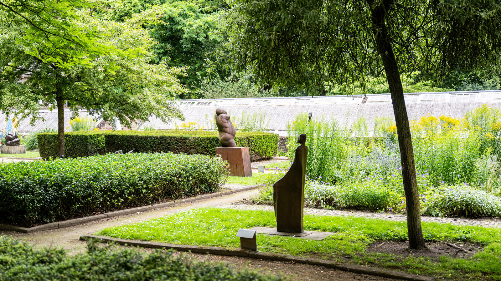 Een vergezicht van een tuin met hagen in de beeldentuin. Twee kleine sculpturen staan op het gras, omringd met bomen.